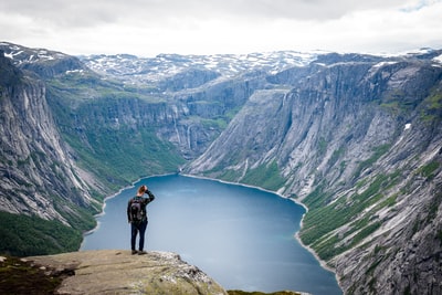 男子在黑色夹克黑色背包站在悬崖上方的一个湖在白天
